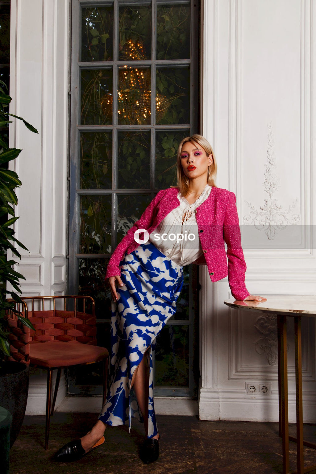 Woman in pink cardigan and blue and white floral pants standing beside window