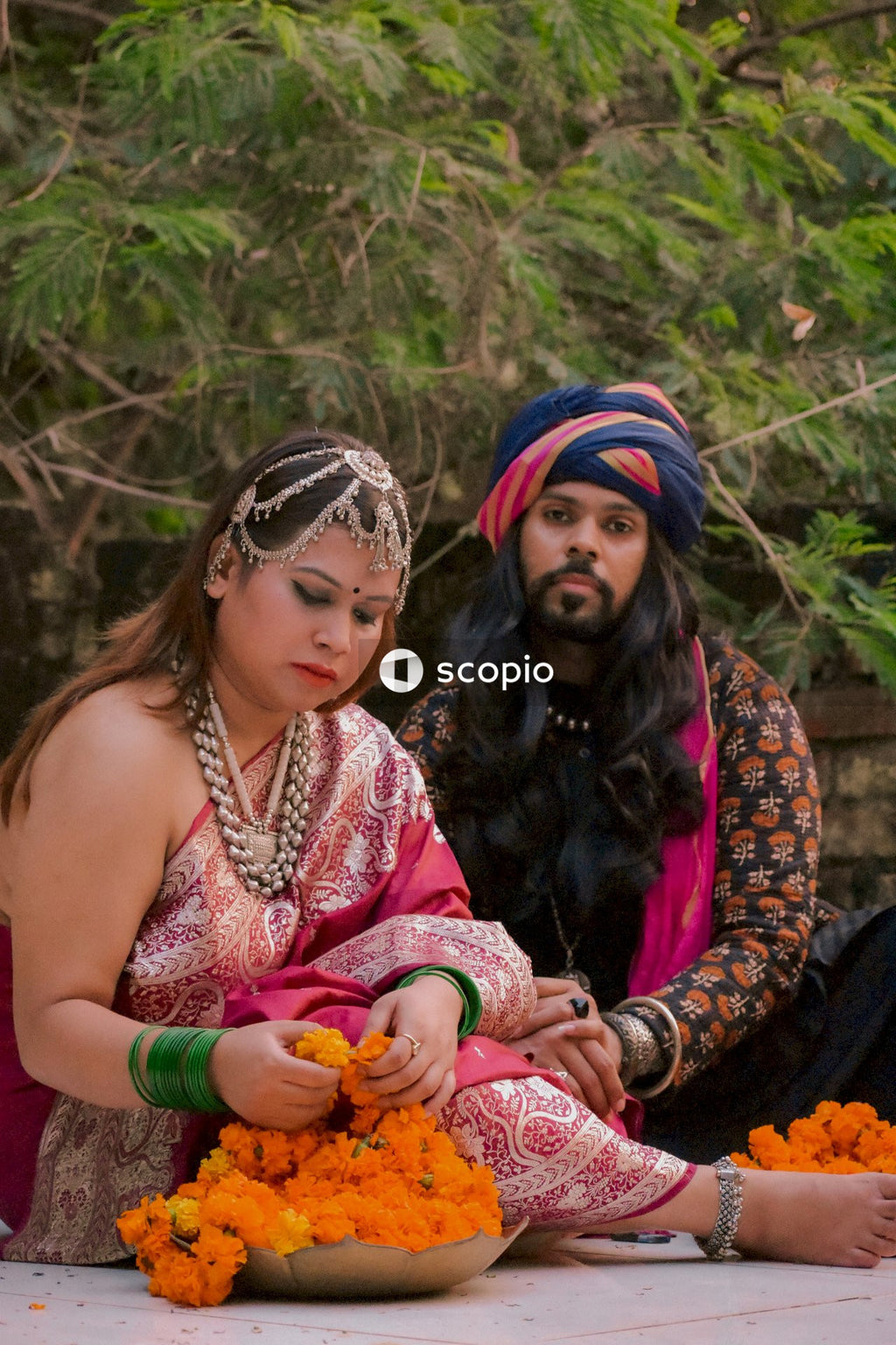 Man with long hair wearing a turban sitting beside woman in pink sari on ground beside trees
