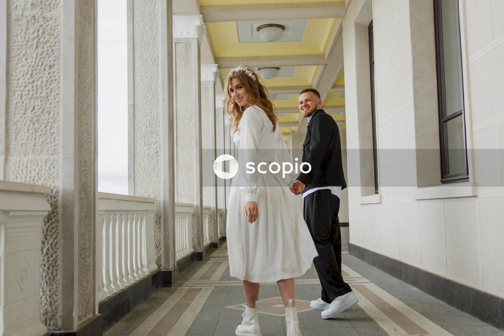 Brunette woman in white long sleeve dress standing beside man in black blazer in a hallway