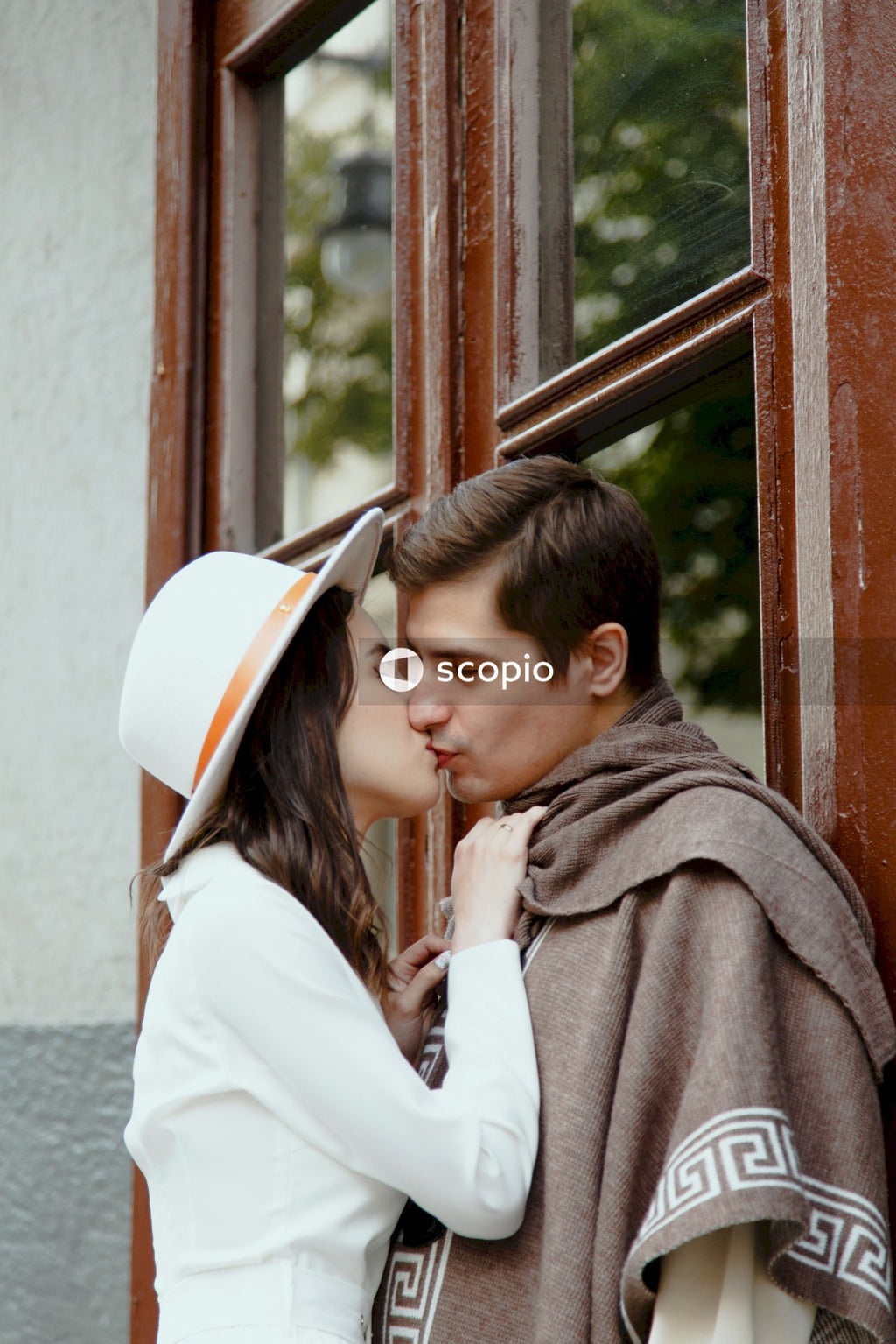 Brunette woman in big white hat kissing Brunette man in beige poncho