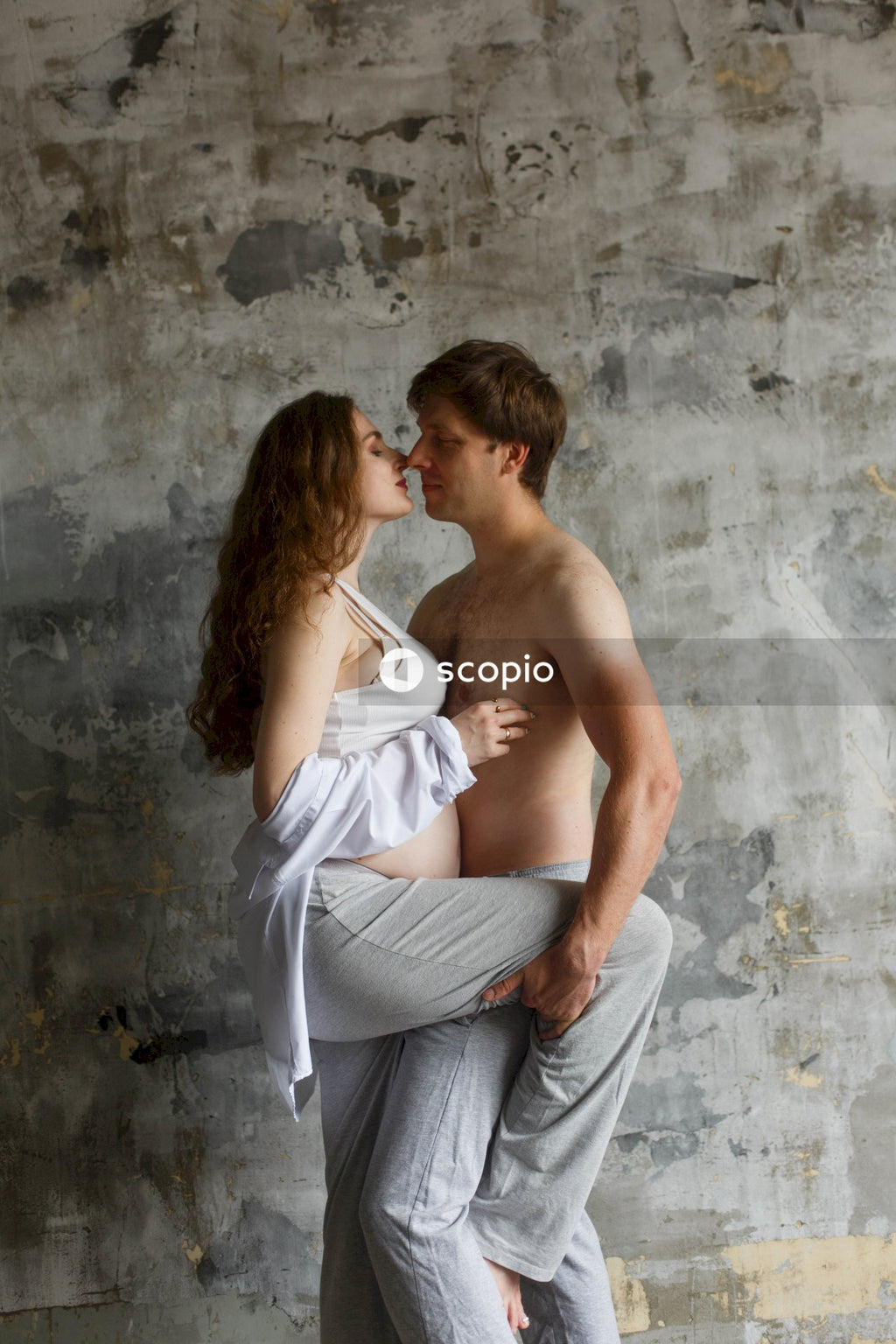 A pregnant  woman in a white tube dress hugging a man in a gray pants