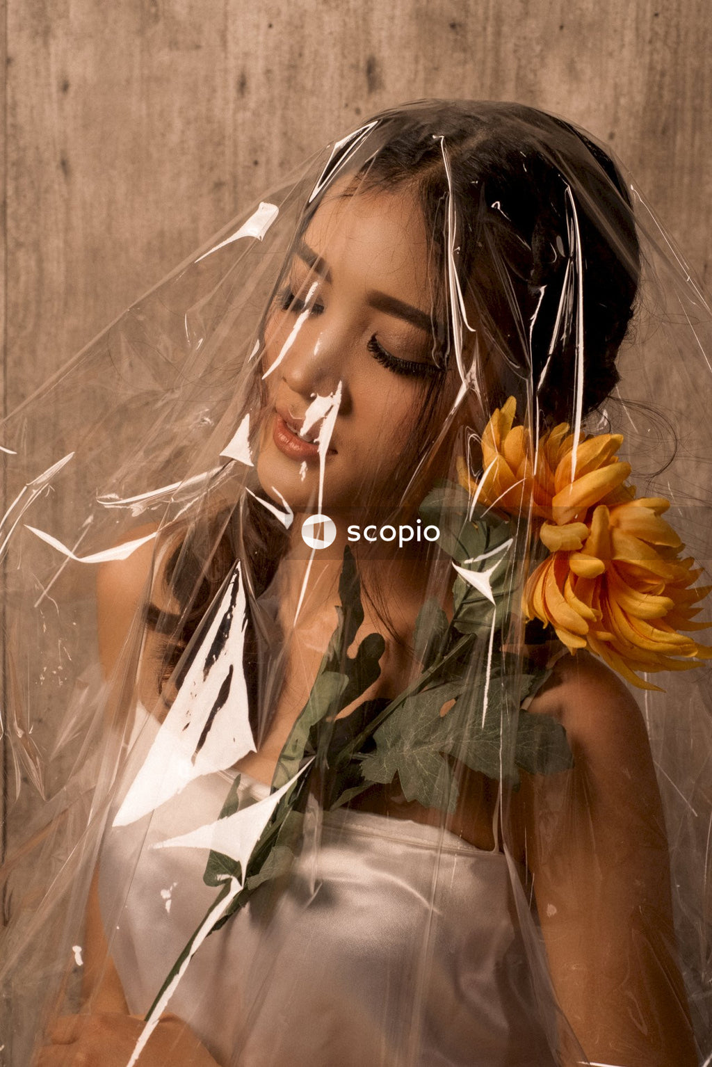 Woman with eyes closed holding yellow flower on her shoulder is covered with cellophane paper