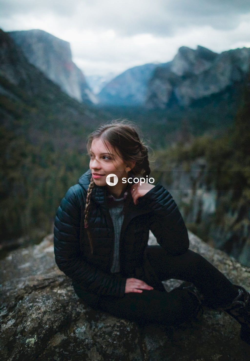 Young woman in dark bubble jacket sitting on rock formation in the woods looking away