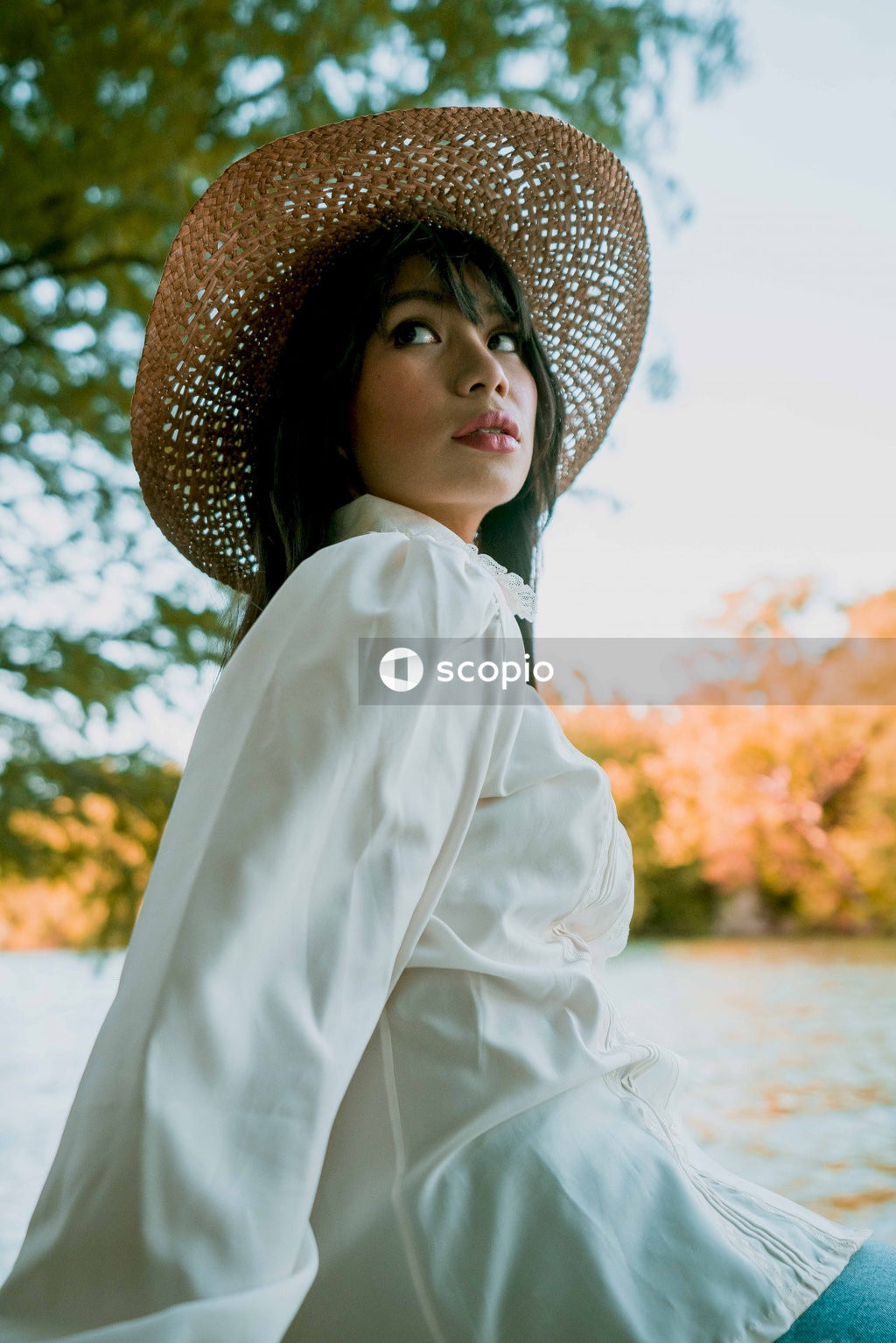 Young woman in light dress wearing straw hat sitting near river
